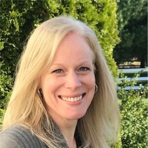 A close up photo of a woman with blonde hair smiling with trees in the background.