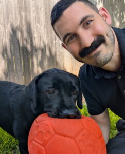 A middle age man with a mustache posed with a black lab puppy that is holding a red soccer ball in his mouth.