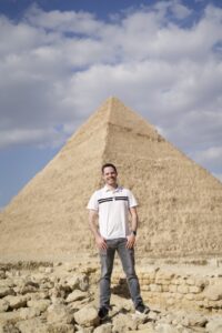 A man stands in the foreground, with an Egyptian pyramid in the background.