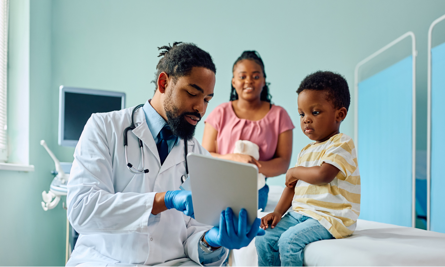 A doctor shows his clipboard to a toddler boy, who is sitting on the examination table, with his mom standing behind him.